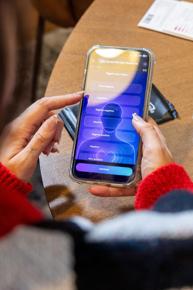 A person with long nails wearing a red sweater is using a smartphone app to select their current mood from options ranging from “Very negative” to “Very positive.”